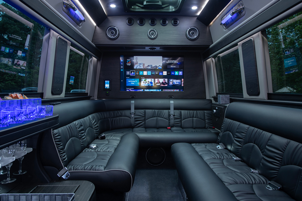 Interior of a sprinter van limousine withe comfortable bench seating, a tv, glasses for drinks and speakers.